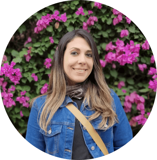 Portrait of Luciana Nunes. She's a brazilian brunette woman with dark medium straight hair bleached in the end. She's wearing a blue jeans jacket. She's in front of wall full of purple flowers.